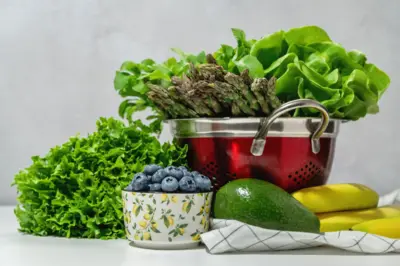 Masada süzgeç içinde taze marul, kuşkonmaz ve roka salatası, mutfak havlusu. Yaban mersini kasesi, muz ve avokado.