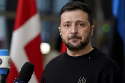 Zelenskyroleta de dinheirofrente a microfones. Ao fundo, há bandeiras e um ambiente desfocado. Ele usa uma camisa preta com um pequeno símbolo no lado esquerdo.