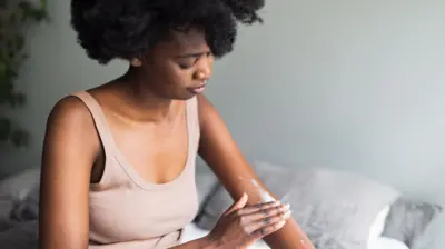A woman sits on a bed and rubs cream into her forearm, looking down as she does this. 