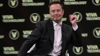 Илон Маск в черном пиджаке и светло-розовой рубашке с расстегнутой верхней пуговицей жестикулирует, выступая перед публикой на черно-белом фоне с логотипом выставки Viva Technology