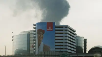 Smoke rising from an area near Dubai International Airport is seen through a vehicle’s windshield after a drone attack hit a fuel tank. In the foreground, an Emirates poster portraying a female flight attendant covers almost the entire façade of a building