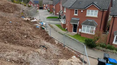 A landslip on a new-build estate. A pile of soil and rubble lies across the road, right up to the pavement on the right. A series of metal barriers edge the landslip. with homes just to the right