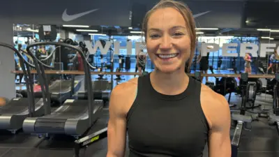 Mulher sorrindo. Ela usa roupas de ginástica e está na academia