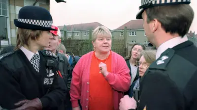Mags Haney frente a su casa en Raploch hablando con dos policías. La foto, de mediados de los 90, muestra a Haney con el pelo corto y rubio decolorado y grandes pendientes. Lleva un cárdigan rosa y una camiseta naranja. Varios vecinos observan la escena.