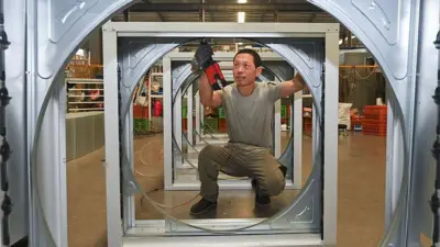 An employee at a wind turbiner manufacturing company uses work tools to put together some steel parts. In the image, he is pictured squatting at the centre of one of the steel frames. 