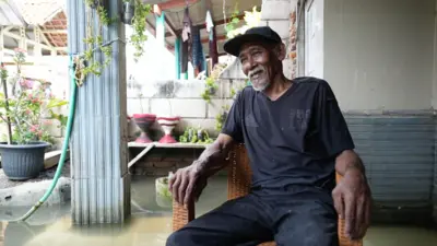 Suwandi está sentado em uma cadeira dentro de sua casa alagada