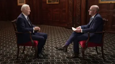 Joe Biden wears a dark blue suit and sits in a red chair in a room with a patterned carpet and wood-paneled walls. He faces the BBC's Nick Robinson, who also wears a dark suit and sits in a chair.