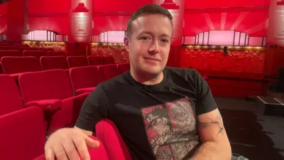 Jack Bence wearing a dark grey T-shirt with anime figures depicted on it sits on red theatre seats in a red room with art Deco-style black and white lights on the walls.