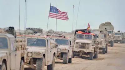 砂道を砂色の軍用車両が荷物を載せ、アメリカ国旗を掲げながら縦一列になり走っている