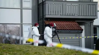 دو تن از افراد فنی طب عدلی که لباس‌های سفید و کلاه سرخ و ماسک آبی دارند، در حال تحقیق در مقابل منزل هستند.