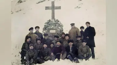 A group of 16 men from the crew of the Quest exploration ship, standing and sitting in front of a memorial cross to Sir Ernest Shackleton