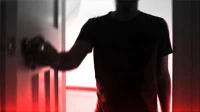 A dark silhouette of a person wearing a short-sleeved shirt is positioned in the foreground, facing a door. The person is using their left hand to grasp and turn a doorknob. The background is brightly illuminated, casting the figure into deep shadow and creating a high-contrast effect. A red gradient overlays the bottom portion of the image, contributing to an eerie or dramatic atmosphere