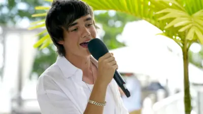 Liam Payne wearing a white shirt and a gold bracelet, holds a microphone and sings on The X Factor 