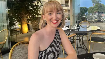 Billie Nicholls in a black and white top sitting at table on holiday. She has blonde shoulder-length hair and a fringe. She wears red lipstick.