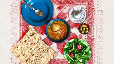 نان سنگک و آبگوشت، سبزی خوردن و زیتون روی یک رومیزی قرمز در کنار گوشت کوب و ظرف خالی و یک قاشق.