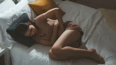 Kobieta leży na łóżku. Trzyma się za brzuch, wygląda, jakby cierpiała.