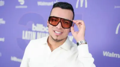 Yeison Jiménez posa ante la cámara llevándose la mano izquierda a las gafas delante de un fondo morado.