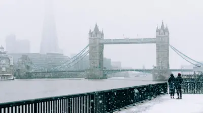 The River Thames and Tower Bridge in London on a snowy day. In the bottom right corner there are two people dressed in dark-coloured coats and hats looking at the view behind black railings.