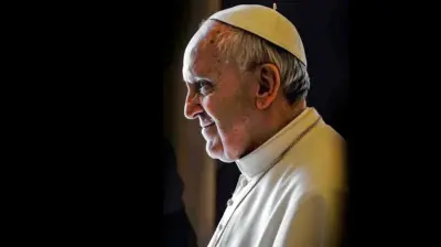 Pope Francis is shown in a head a shoulders shot. He is wearing a white coat and white cap. He is shown in profile looking off the the left.