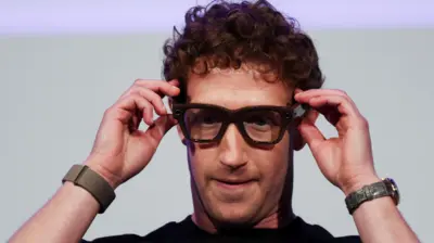 O CEO da Meta, Mark Zuckerberg, em um palco, gesticulando em direção aos óculos de armação grossa em seu rosto, que são um protótipo dos óculos inteligentes alimentados por IA ainda não lançados pela empresa. Ele veste uma camiseta preta e um relógio de pulso.
