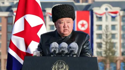 Kim Jong-un habla desde un atril con tres micros. Lleva un gorro y tiene la bandera de su país detrás.