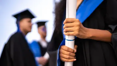 Elinde rulo halinde diploma tutan üniversite mezuniyet kıyafeti giyen bir kadın ve arka planda mezuniyet kepli iki erkek öğrenci