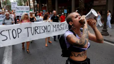 A demonstrator with tattoos uses a megaphone as people protest against mass tourism on a street in Alicante, Spain, July 13, 2024. The banner reads "Stop turismo masivo" ("Stop mass tourism")