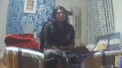 Imagens de câmera oculta mostram uma mulher asiática sentada em uma cama. Parede com papel de parede azul vivo e prata ao fundo, enquanto a colcha apresenta um padrão xadrez grande em amarelo, azul e cinza