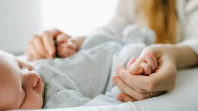 Yeni doğan bir bebek sırt üstü uzanırken, sarışın bir anne bebeğin ellerinden tutuyor