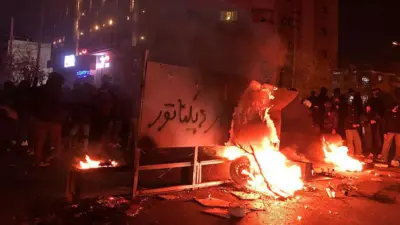 آتش روشن است و بر یک تابلوی شهری نوشته شده است مرگ بر دیکتاتور. جمعیت اطراف ایستادهاند و به نظر میرسد نردههای خیابان کنده شده است.