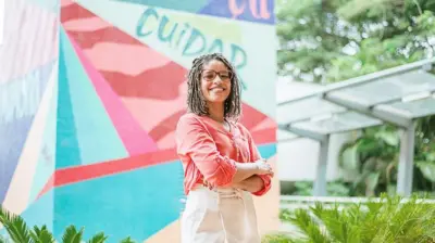 Mariene Ramos posando para a foto sorrindo. Ela tem os cabelos trançados em estilo afro e usa óculos escuros, camisa salmão e calças brancas. Está com os braços cruzados sobre o peito e olha para a câmera meio de perfil. Ao fundo se vê um mural colorido com a palavra "cuidar" em destaque