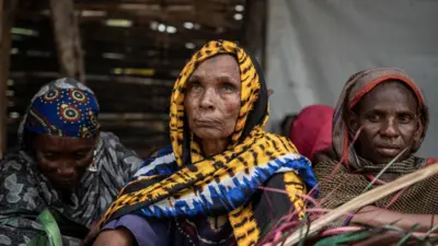 Yaşlı bir kadın yukarı bakarak birini dinliyor. Yanında iki kişi daha var.