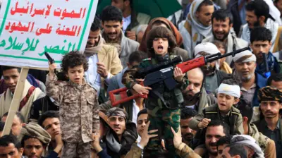 Толпа на митинге. В центре на плечах взрослых двое детей в камуфляже с макетами пистолета и автомата