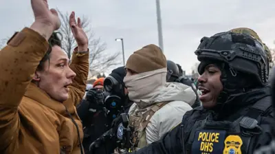 A demonstrator in a brown jacket holds their arms above their head in front of a uniformed official