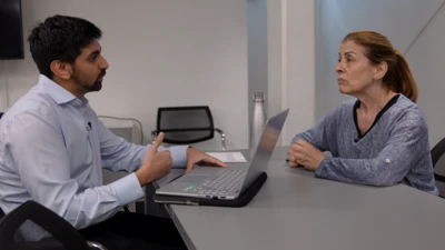 A man and a woman sit across a grey table from each other. The man, on the left, has a laptop open in front of him and is talking while gesturing with his right hand. The woman, on the right, is looking at him, listening.