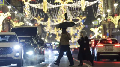 Двое людей переходят улицу, украшенную новогодними гирляндами