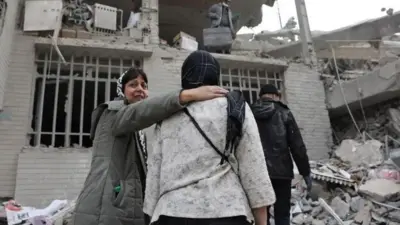 یک ساختمان ویران شده و چند نفر در اطراف آن، یک زن ناراحت دستش روی شانه زن دیگری انداخته است