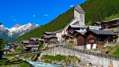 A general view of a row of buildings in Blatten, on an elevated road overlooking a stream. There are forest-covered mountains behind. The sky is bright blue and there are snow-topped mountains in the background.