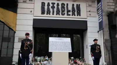 「BATACLAN(バタクラン)」という看板が据えられたホールの玄関前に、男女の衛兵が立っている。玄関前には事件の犠牲者の名前が書かれたボードが置かれている。玄関のガラスのドアには、献花の花束が山と積まれている