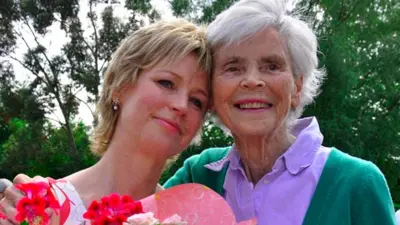 Sally Magnusson (left), holding red and pink flowers, with her mother Mamie who is wearing a purple blouse and green cardigan. They are looking in the same direction and their heads are touching. There are green trees behind them. 