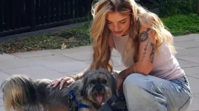 Giuliana Passarelli este surprinsă în timp ce se apleacă și mângâie un câine. Are părul lung și blond și poartă un maiou alb fără mâneci, care îi lasă la vedere tatuajele de pe brațul stâng.