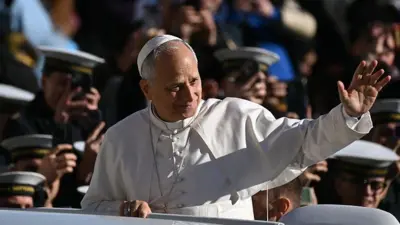 El Papa, vestido de blanco, saluda a la multitud desde su papamóvil con un gesto de la mano izquierda. Hombres o niños uniformados se toman fotos al fondo.