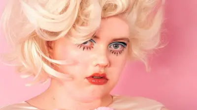 Megan Winstone looking directly into the camera. She is in exaggerated eye make-up and has large blonde curly hair.