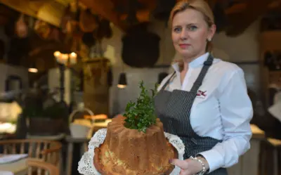 Kobieta stojąca w rustykalnym wnętrzu restauracyjnym trzyma na wyciągniętych dłoniach dużą, domową babkę wielkanocną ustawioną na białej, koronkowej serwetce. Wypiek ma kształt wysokiej formy z kominem, a na jego wierzchu znajduje się świeża zielona gałązka. 