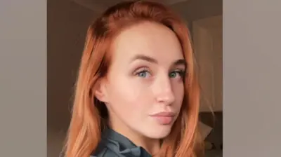 Uma mulher de cabelo ruivo e olhos azuis sorrindo.