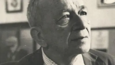A imagem em preto e branco mostra um retrato em close de um homem idoso, Mario Quintana, de cabeça parcialmente calva, usando terno e camisa social. Ele aparece com o olhar voltado para cima e para a direita, em expressão serena e reflexiva, enquanto o fundo desfocado revela quadros pendurados na parede e outra pessoa sentada ao longe.