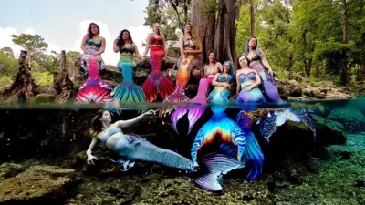 Grupo de mulheres vestidas com caudas de sereia posa para foto à beira d'água