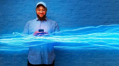Un homme souriant, son téléphone portable à la main, se tient devant un mur de briques peint en bleu, orné d'une illustration de rayons de lumière bleue le traversant.