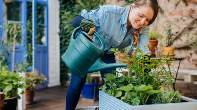 A woman in a denim top waters potted plants and smiles while looking at them. There are other potted plants in the background, while she stands on her patio.