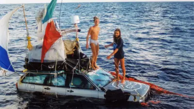 Dos hombres —Marco Amoretti y Marco De Candia— están de pie sobre el capó de un coche que flota en el océano. Al coche le cuelgan redes rojas, cuerdas y banderas. Llevan las piernas al descubierto, y uno de ellos está desnudo, salvo por unos calzoncillos blancos. El otro lleva una camiseta azul marino. Tienen barba y pelo largos, y son delgados y morenos.